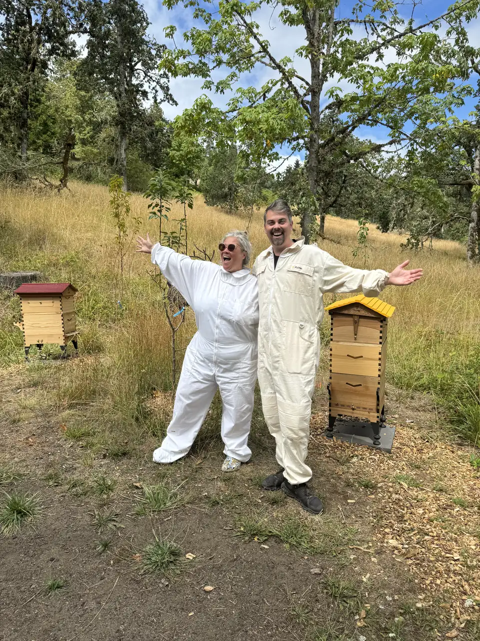 Kathryn and Geoff Belleville in their bee suits with veils off, arms wide, between the red-roofed and yellow-roofed hives